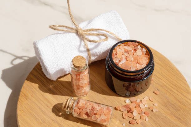 Pink Himalayan salt in the glass bottles with rolled towel on wooden board. Close up of pink crystals of sea salt. Spa body care concept.